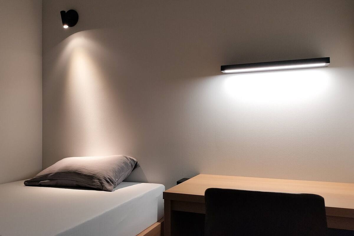 Quarto com uma cama, uma escrivaninha e luminária fixada na parede.