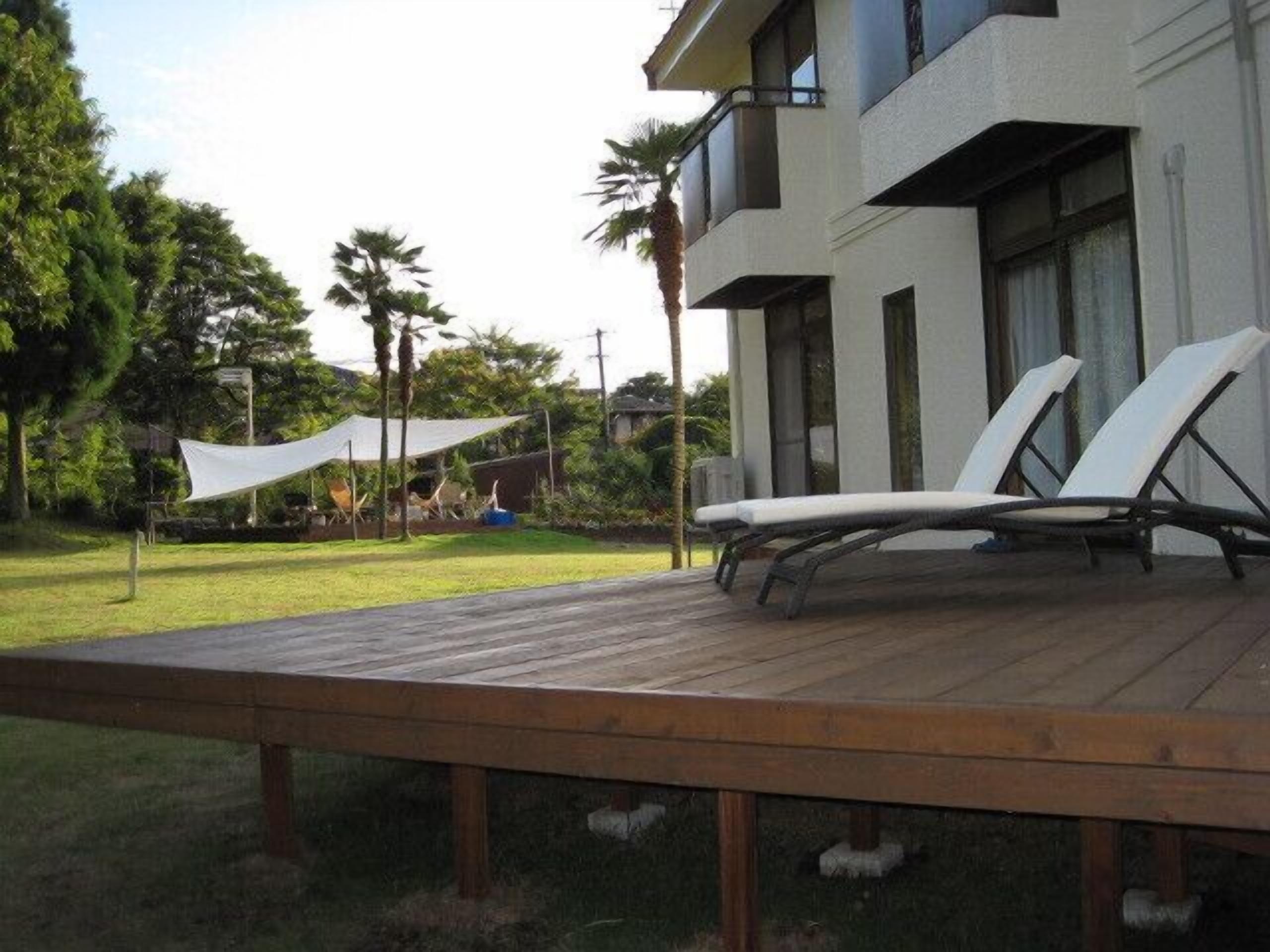 Una terraza de madera con dos sillones de descanso, un dosel blanco y un edificio moderno con ventanas y balcones amplios.