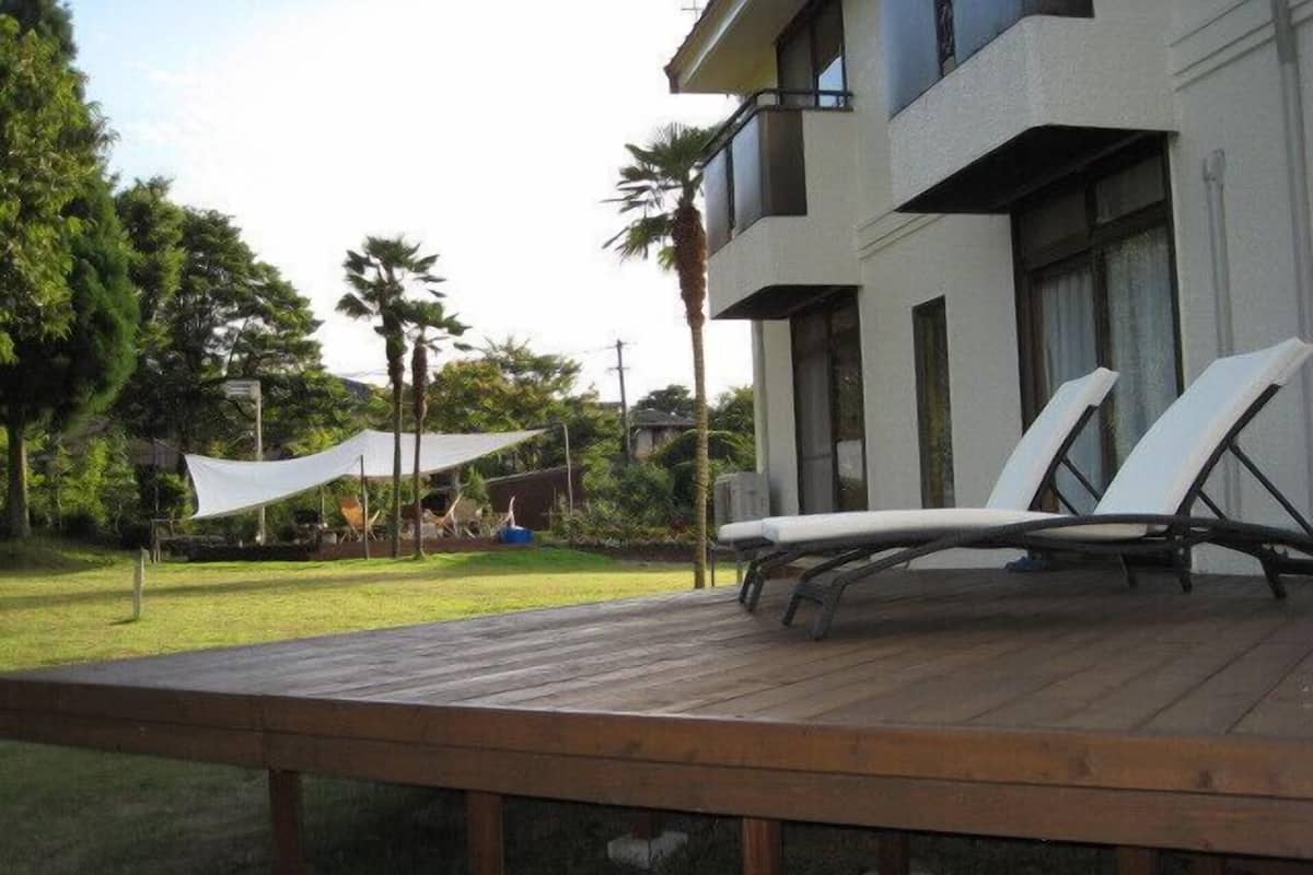 Een houten terras met twee loungestoelen, een wit zonnescherm en een modern gebouw met grote ramen en balkons.
