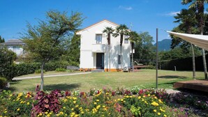 Una casa blanca de dos pisos amb un teulat vermell, envoltada per un jardí ben cuidat ple de flors colorades i palmeres.
