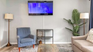 A living room with a flat-screen TV mounted on the wall, a blue armchair, a wooden coffee table, a grey side table with decorative items, a beige sofa, and a tall floor lamp. - Pool, Fenced Yard, & Patio! Ventura Vista Retreat (Columbus)