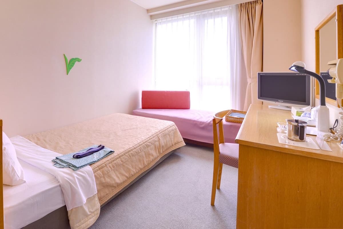 Una camera d'albergo con un letto, una scrivania con un computer, una sedia, una lampada da scrivania, un telefono e una finestra con tende.