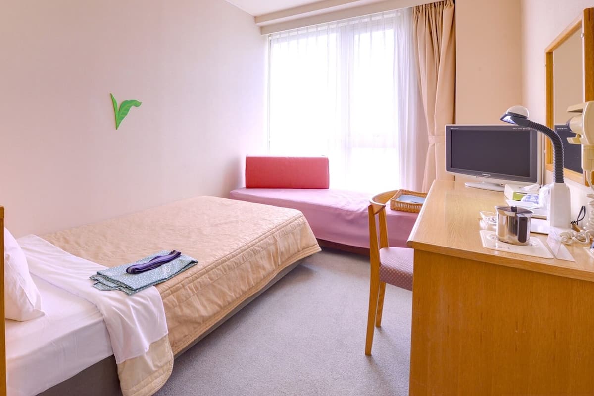 Una camera d'albergo con un letto, una scrivania con un computer, una sedia, una lampada da scrivania, un telefono e una finestra con tende.