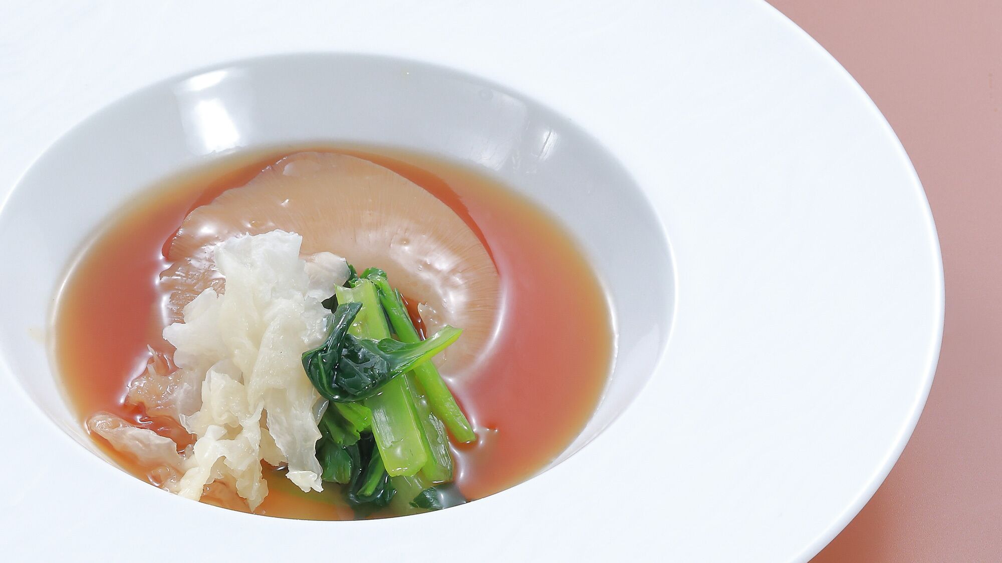 Een witte schaal met soep, bestaande uit heldere bouillon, witte noedels en groene groenten.
