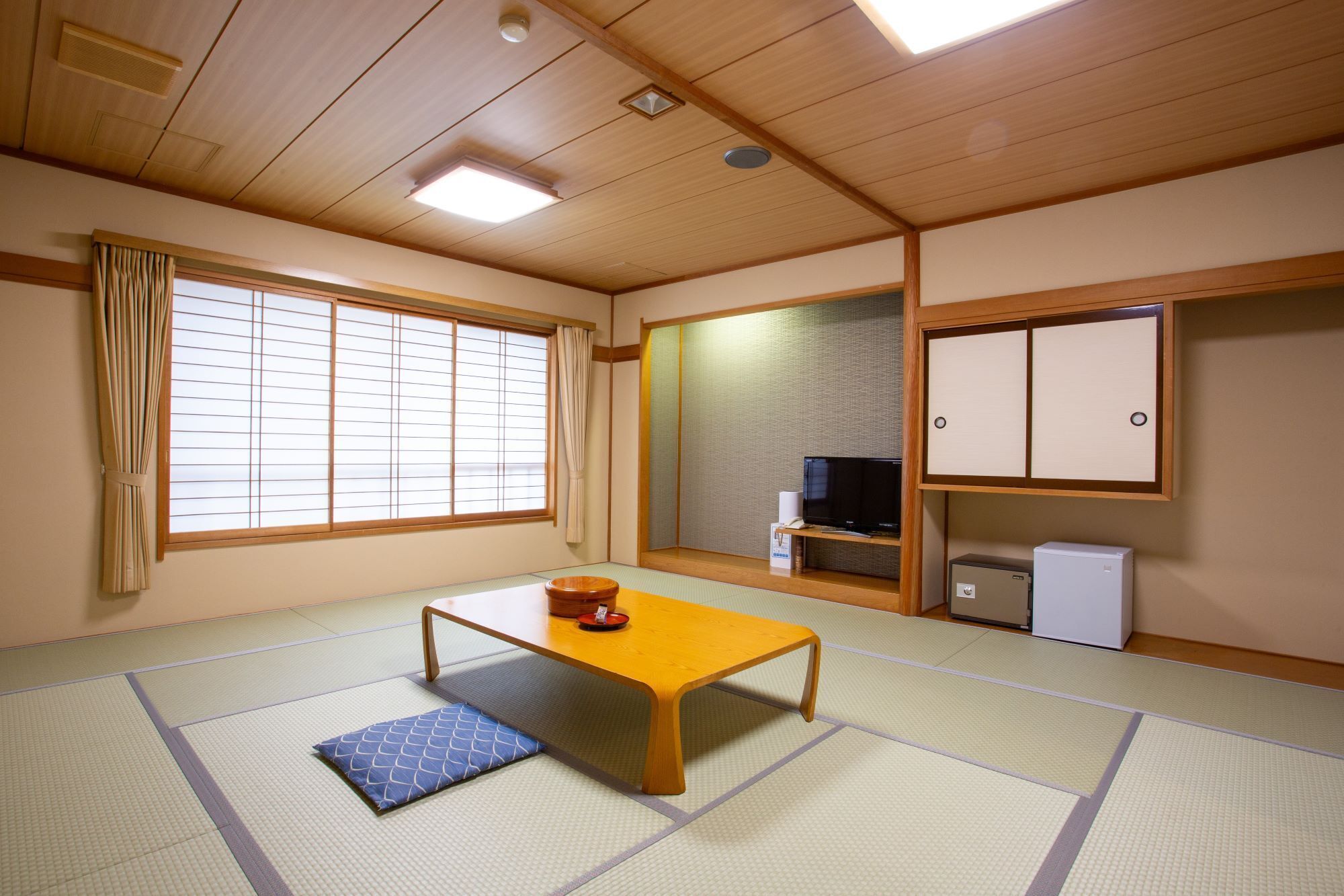 Una habitación con esterillas de tatami, una mesa de madera baja, un televisor y un refrigerador pequeño.