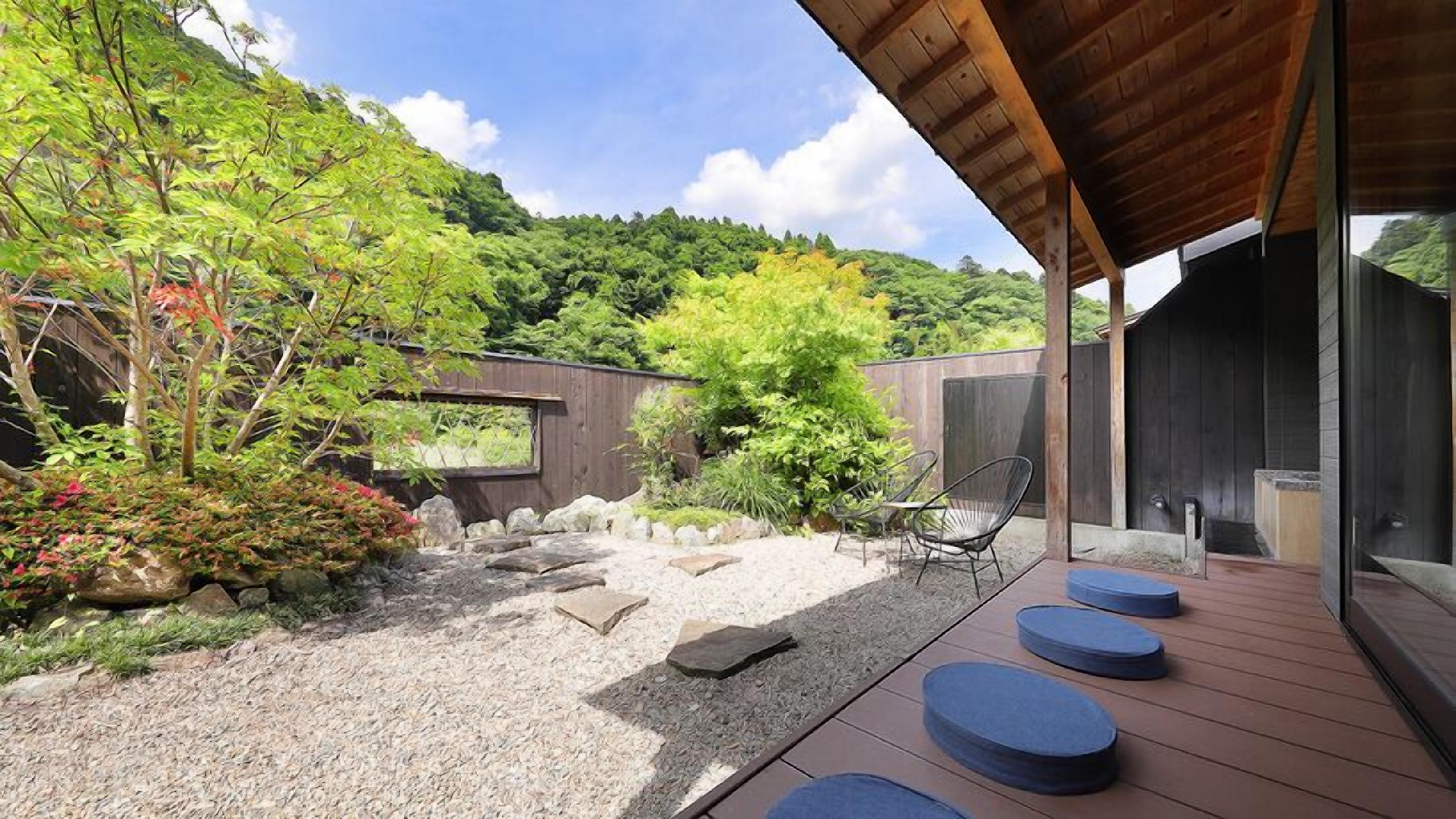 A modern outdoor patio with a wooden deck, seating, and a view of a lush garden and hillside.