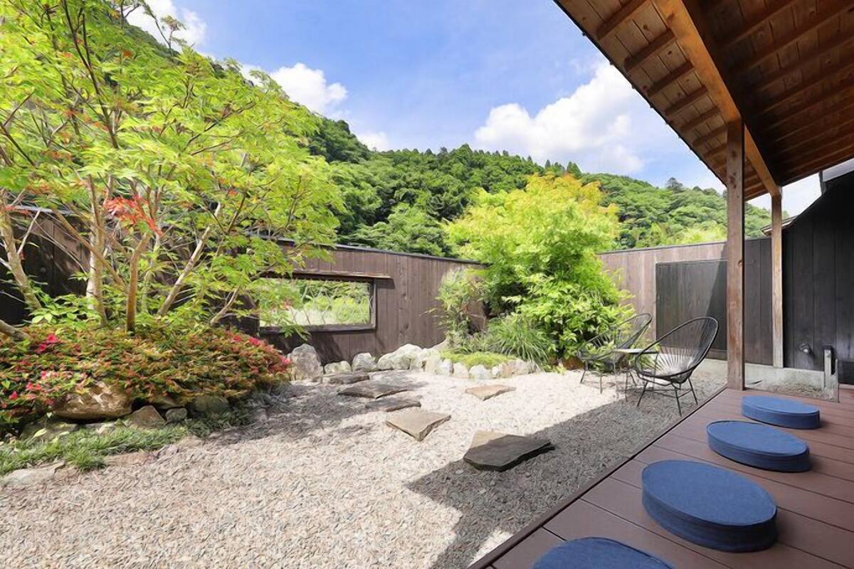 A modern outdoor patio with a wooden deck, seating, and a view of a lush garden and hillside.