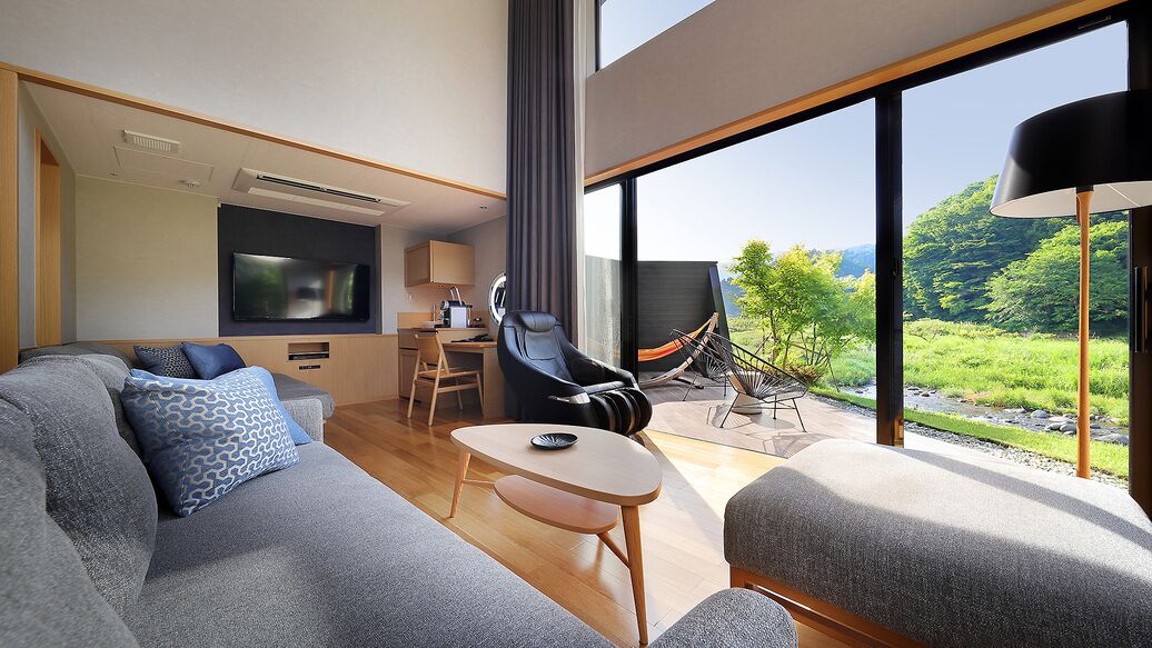 A modern hotel room with a sofa, a small table, a TV, a chair, and a view of greenery outside.