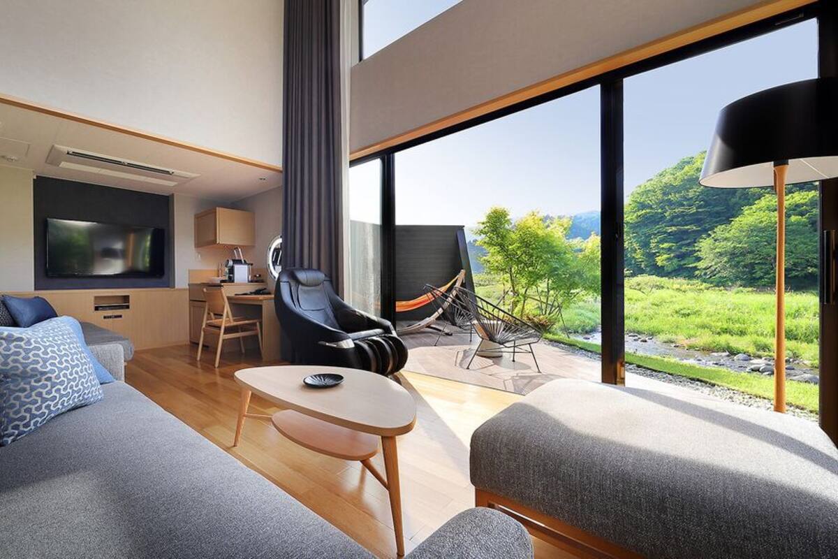 A modern hotel room with a sofa, a small table, a TV, a chair, and a view of greenery outside.