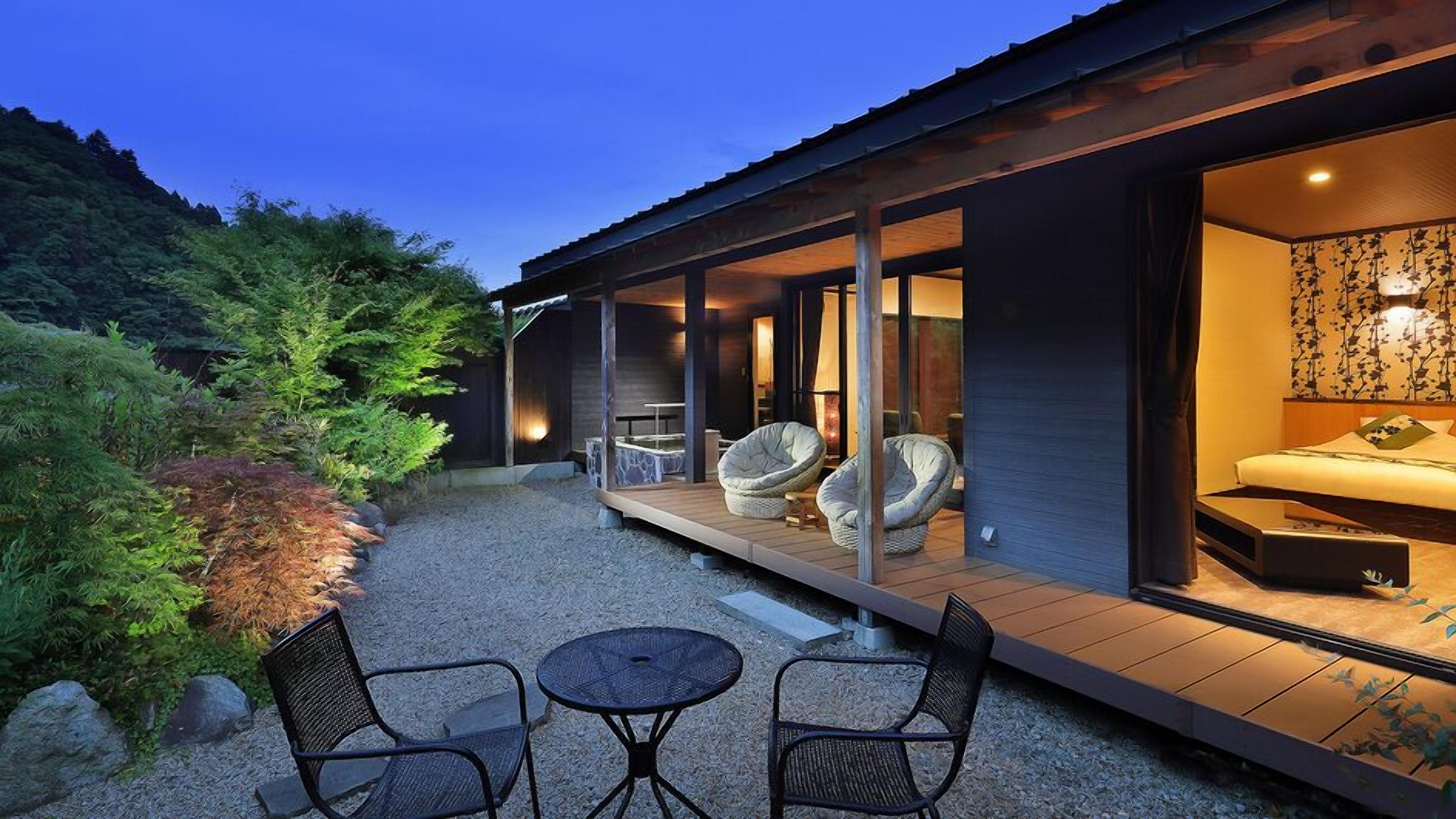 A modern house with a covered patio, outdoor seating, and a bedroom visible through an open door.