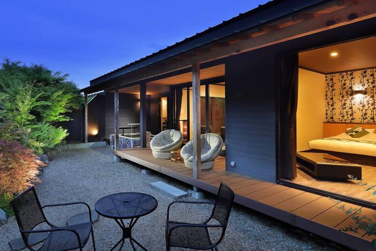 A modern house with a covered patio, outdoor seating, and a bedroom visible through an open door.