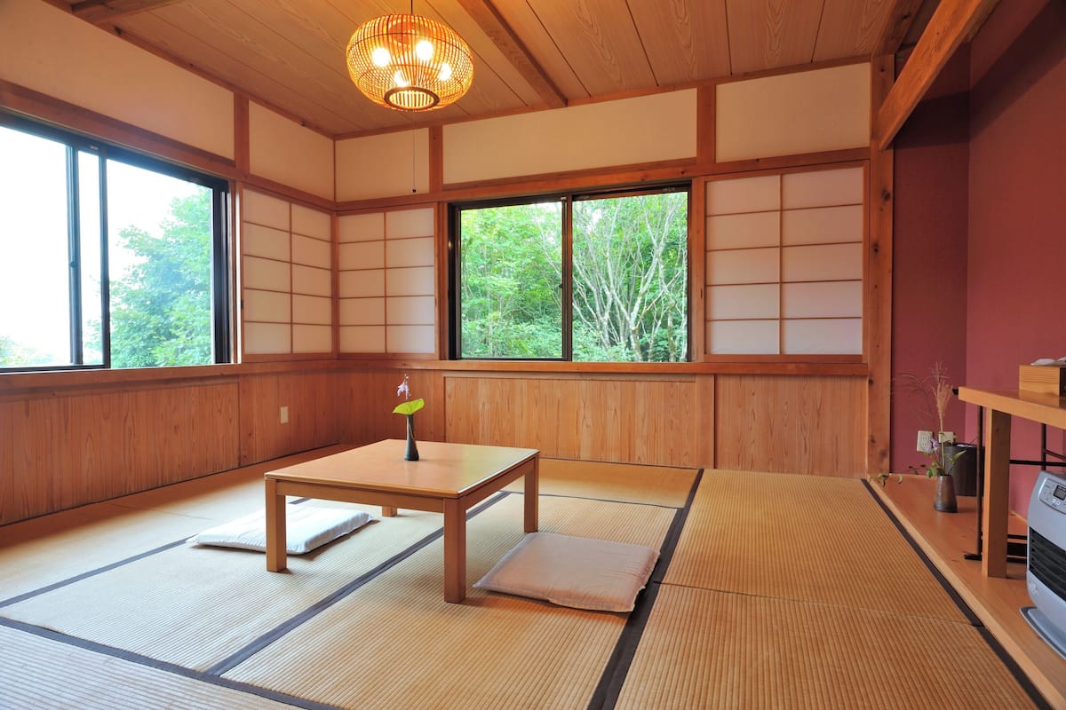 A traditional Japanese room with tatami flooring, a low wooden table, a shoji screen, and a view of greenery outside.