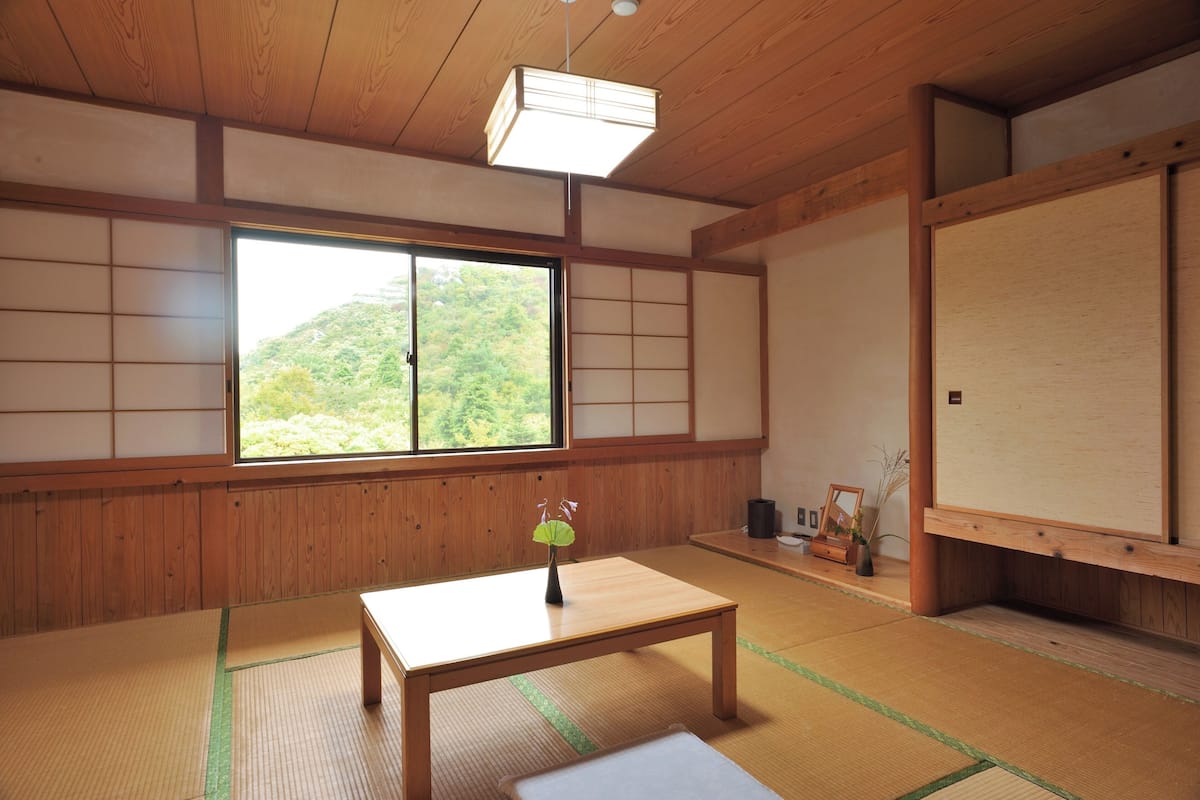 A traditional Japanese room with tatami flooring, a low wooden table, a sliding door, and a view of a green hillside.
