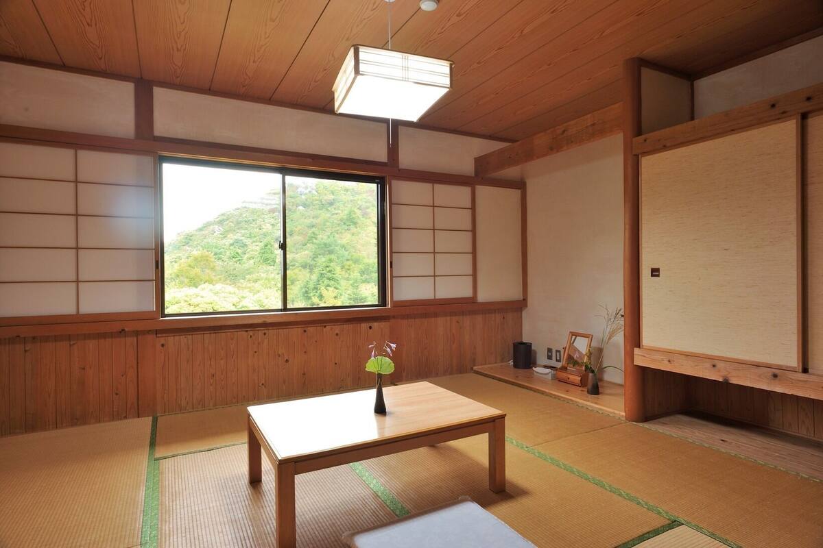 A traditional Japanese room with tatami flooring, a wooden table, a window with a view of greenery, and shoji screens.