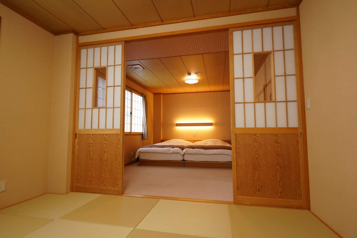 Une pièce traditionnelle japonaise avec un sol recouvert de tatamis, un lit bas en bois, des portes coulissantes en papier de riz (shoji) et un luminaire au plafond.