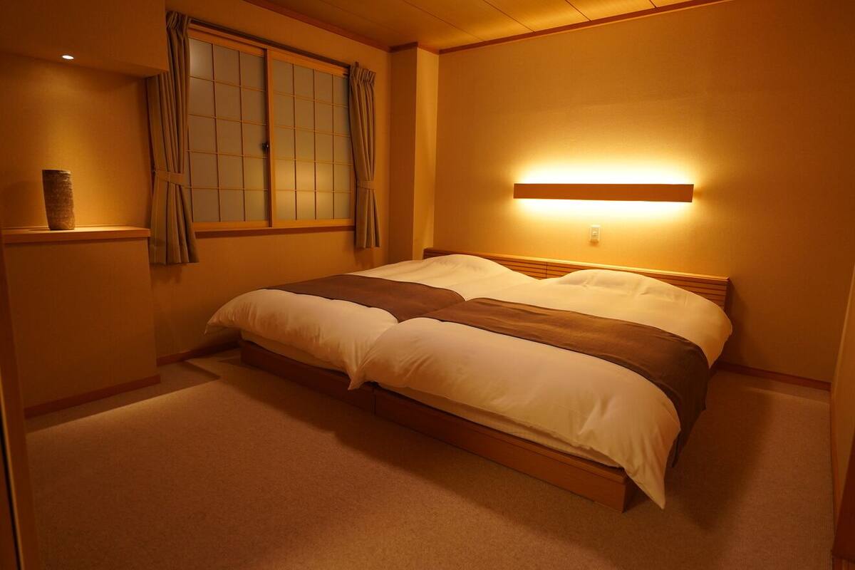 Une chambre d’hôtel avec un lit, une fenêtre avec des rideaux, une table de chevet avec un vase et une lampe fixée au mur.