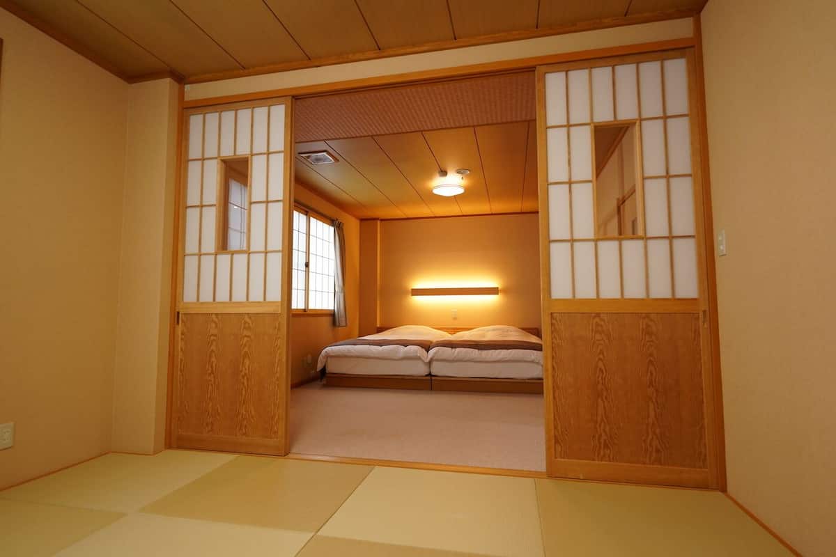 Une pièce de style japonais traditionnel, avec un sol recouvert de tatamis, un lit bas, des portes coulissantes et un luminaire au plafond.