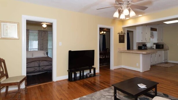 A living room with hardwood flooring, a ceiling fan, a flat-screen TV, a coffee table, and a kitchen area with white cabinets and a countertop. - Mins to Ft Moore & Downtown - The Columbus Cottage (Columbus)