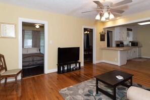 A living room with hardwood flooring, a ceiling fan, a flat-screen TV, a coffee table, and a kitchen area with white cabinets and a countertop. - Mins to Ft Moore & Downtown - The Columbus Cottage (Columbus)