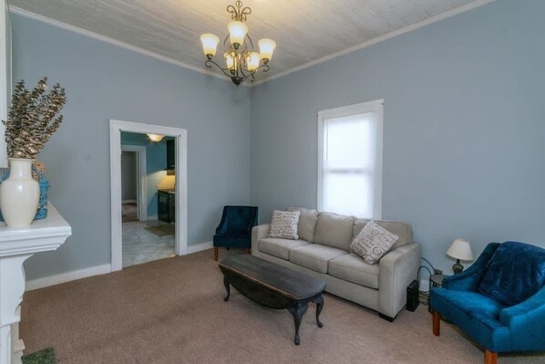 Un salon avec un canapé beige, une table basse en bois foncé et un fauteuil bleu. Sur une console blanche, on aperçoit un vase blanc contenant des plantes séchées. Un lustre est suspendu au plafond et une fenêtre laisse entrer la lumière naturelle.
