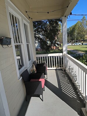 A covered porch with two wicker chairs and a small table, white railings, and a view of a street with cars and trees. - Moments to Chattahoochee-Cottage - Walk-To-Dining! (Columbus)