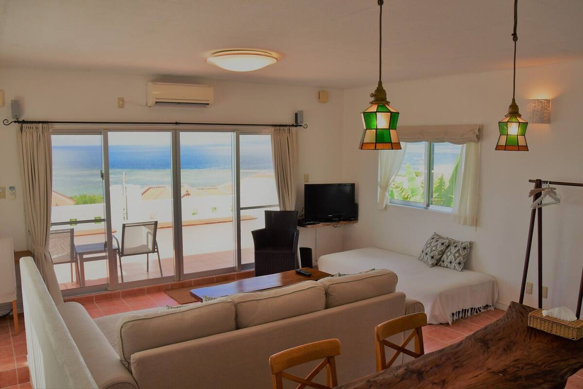 Ein Zimmer mit einer Couch, einem Fernseher, einem Tisch und Meerblick durch große Fenster.