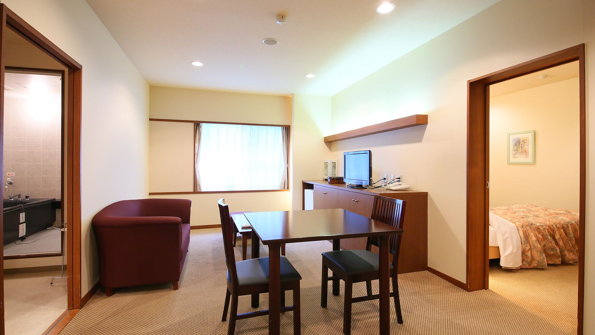 A hotel room with a dining table, four chairs, a red sofa, a bed with a floral bedspread, a desk with a computer and phone, and a window with curtains.