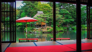Et traditionelt japansk rum med tatamigulv, en rød bænk og udsigt til en dam med en bindingsværksbygning.