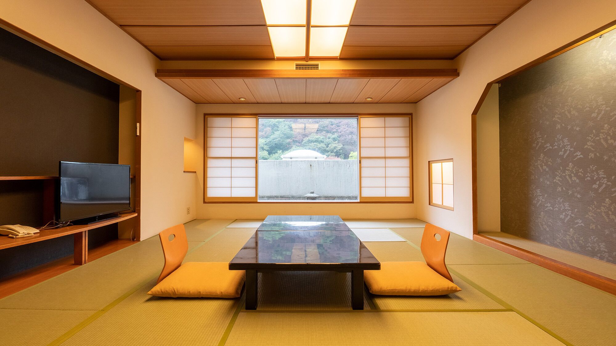 A traditional tatami room with a tatami mat floor, a low table, cushions, a television, and sliding shoji doors.