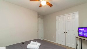 Quarto com uma cama, ventilador de teto, televisão sobre um suporte e uma porta.