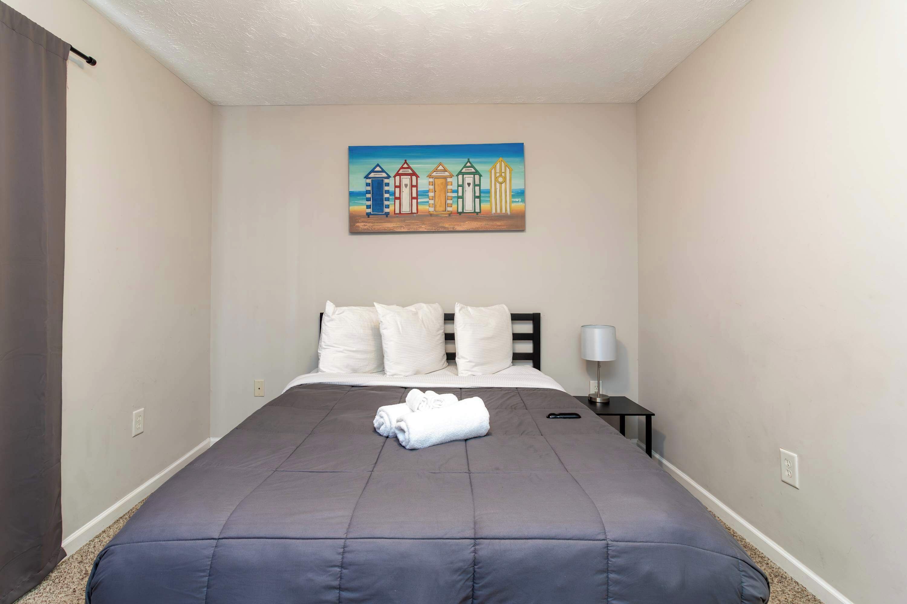 A hotel room with a bed, pillows, a painting of beach huts, a nightstand, and a lamp.