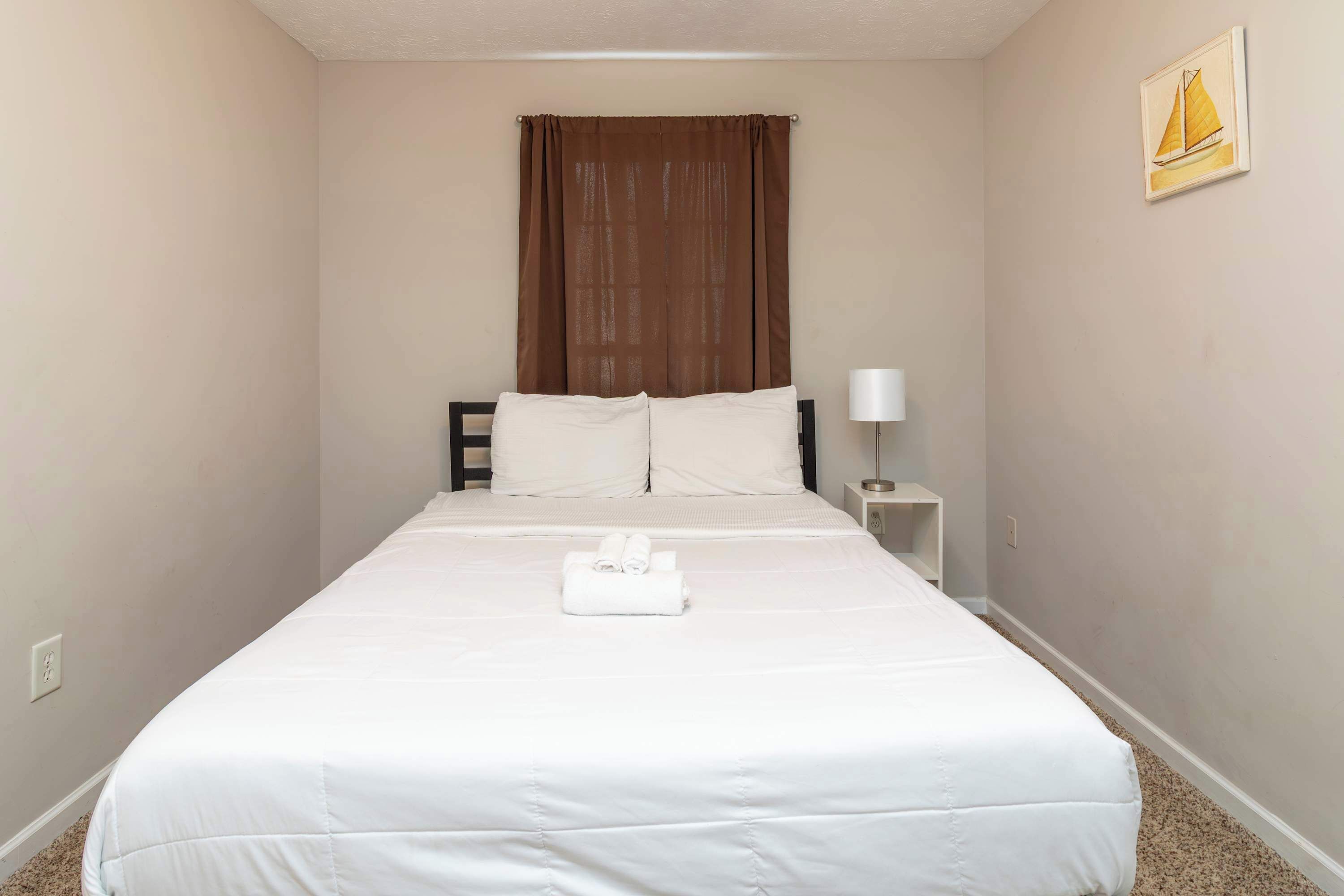 Un lit bien fait, avec du linge de lit blanc, un rideau marron, une petite table de chevet avec une lampe et un tableau encadré au mur.