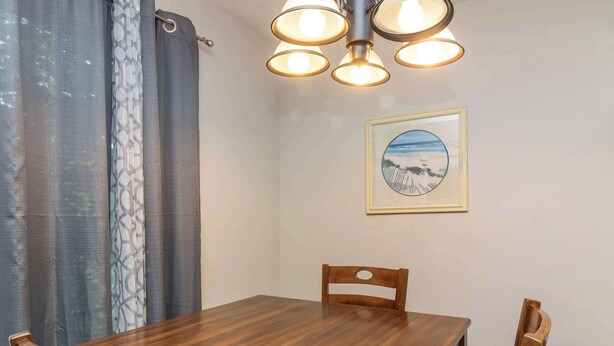 Uma área de refeições com mesa de madeira, cadeiras, luminária no teto e um quadro na parede.