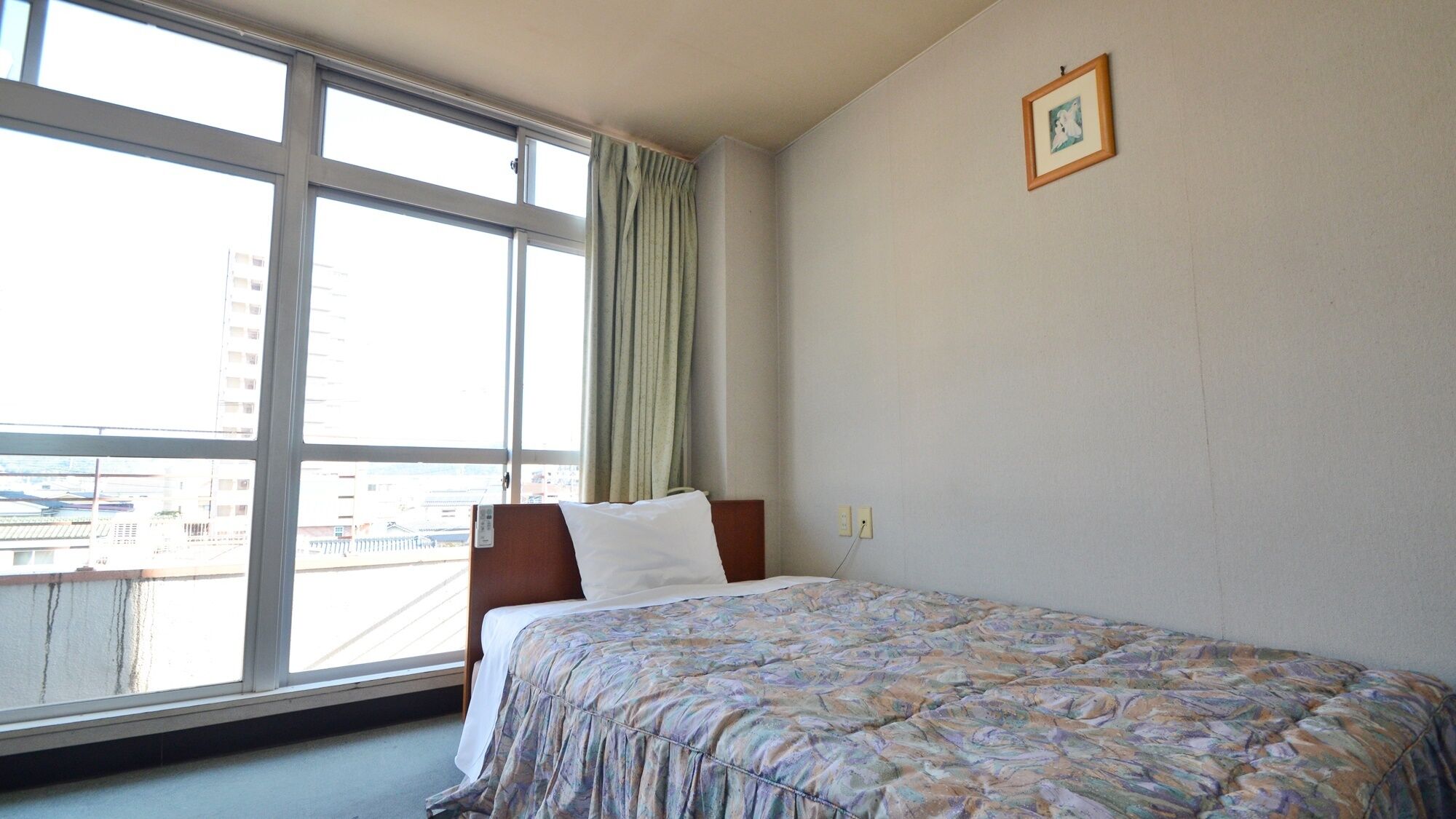 A hotel room with a large window, a bed with a patterned bedspread, a wooden headboard, and a framed picture on the wall.