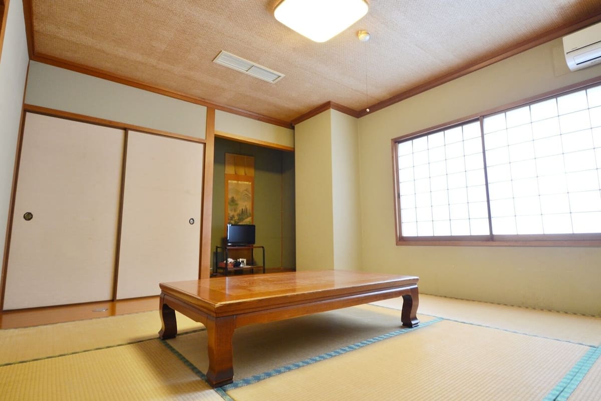 Una habitación japonesa tradicional con suelo de tatami, una mesa de madera baja, puertas correderas y un ventanal grande.