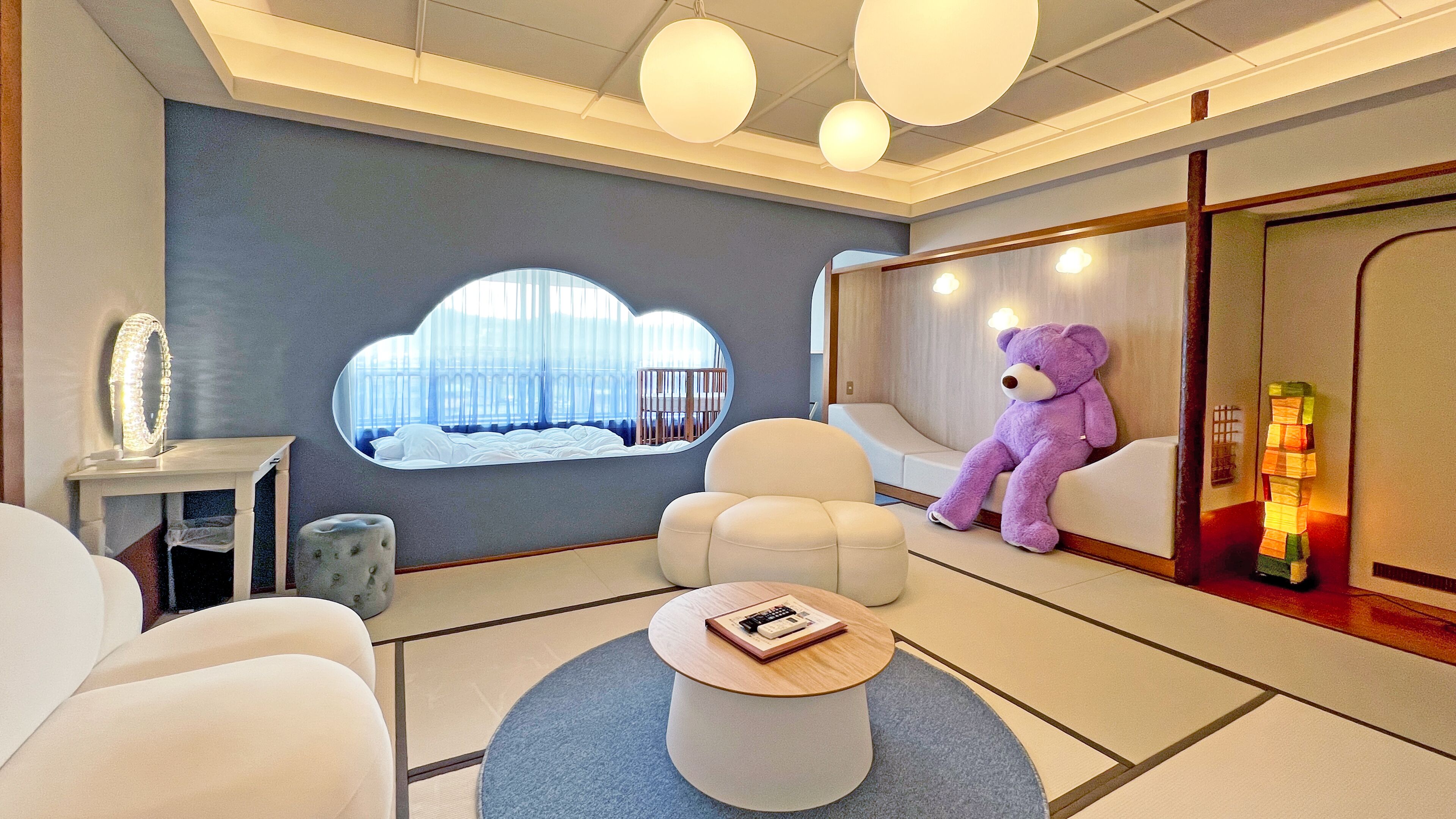 Una habitación de hotel moderna con un ventanal grande, un sofá mullido, una mesa de centro redonda y un osito de peluche.