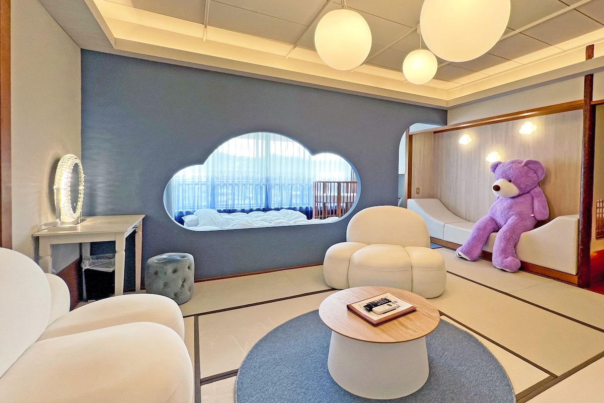 Une chambre d’hôtel moderne avec une grande fenêtre, un canapé moelleux, une table basse ronde et un ours en peluche.