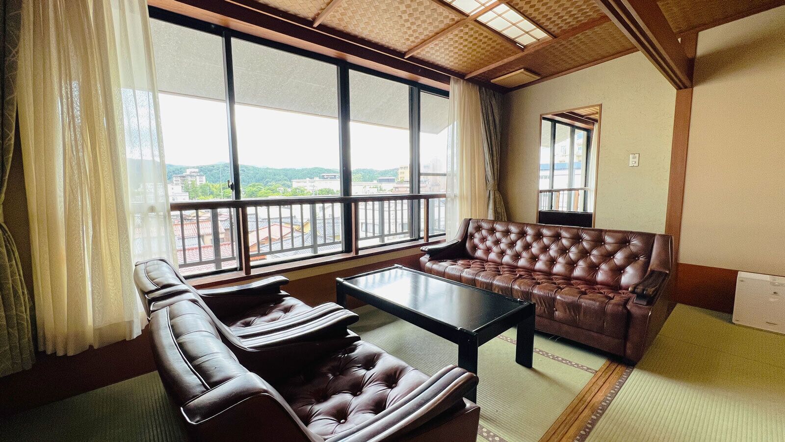 Una habitación con sofá, una silla, una mesa, ventanas amplias y vistas a edificios.