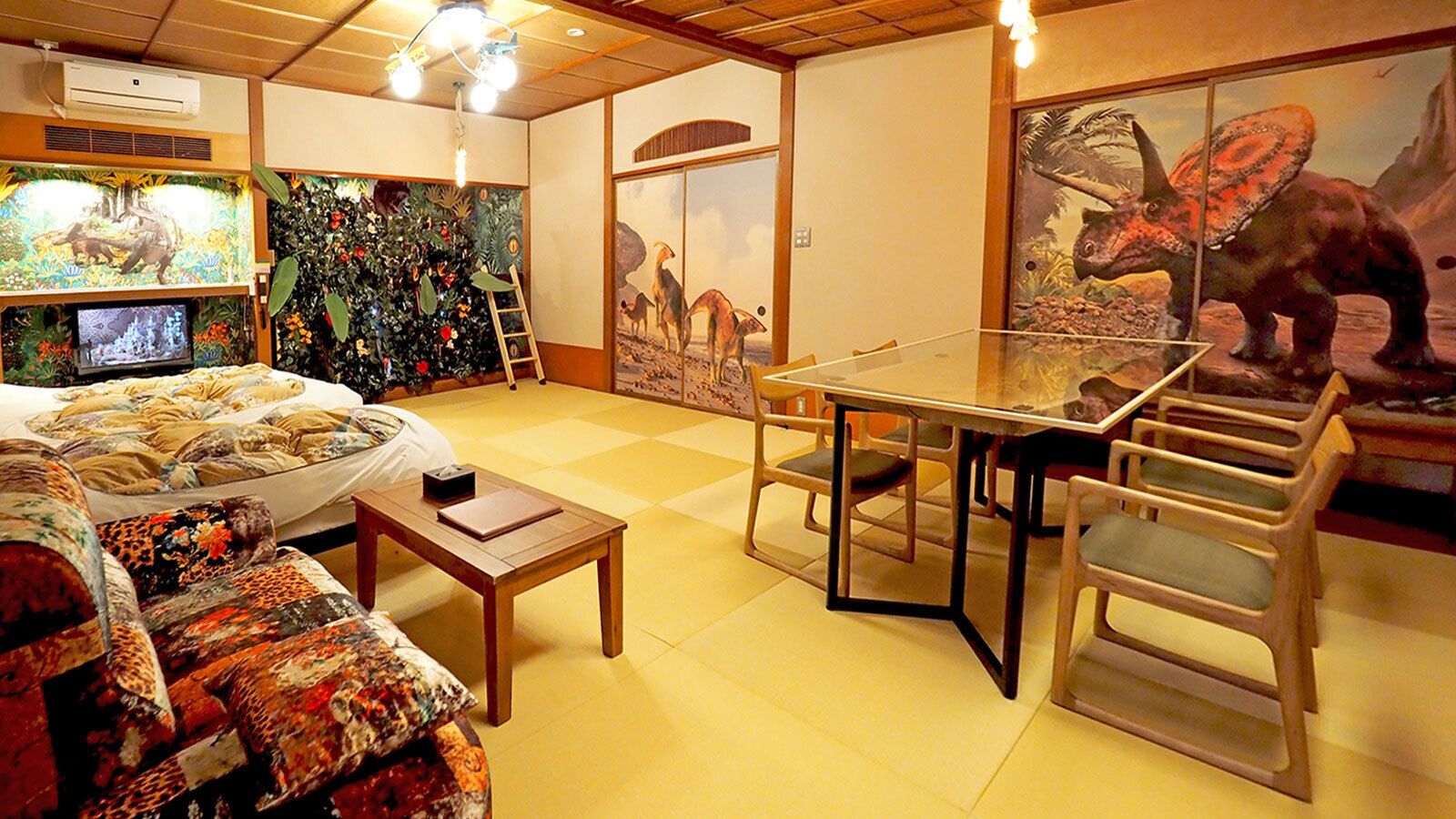 Una habitación con cama, una mesa, sillas, televisor y decoración temática de dinosaurios.
