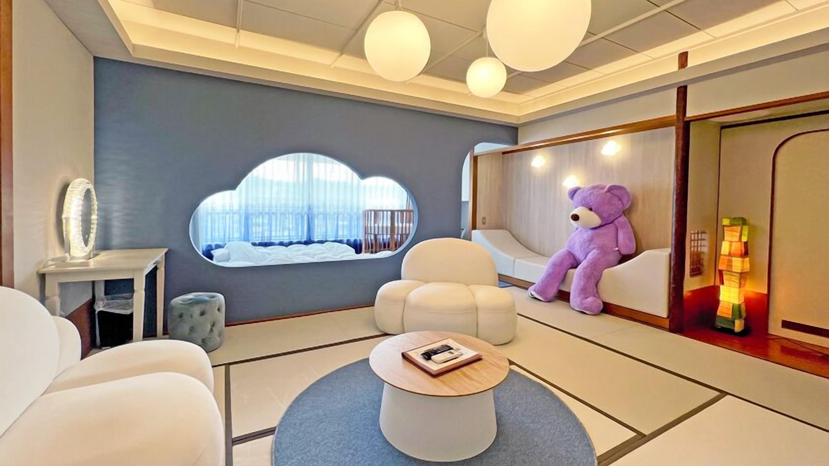 Habitación de hotel moderna con un ventanal grande, un sofá cómodo, una mesita de centro redonda y un osito de peluche.