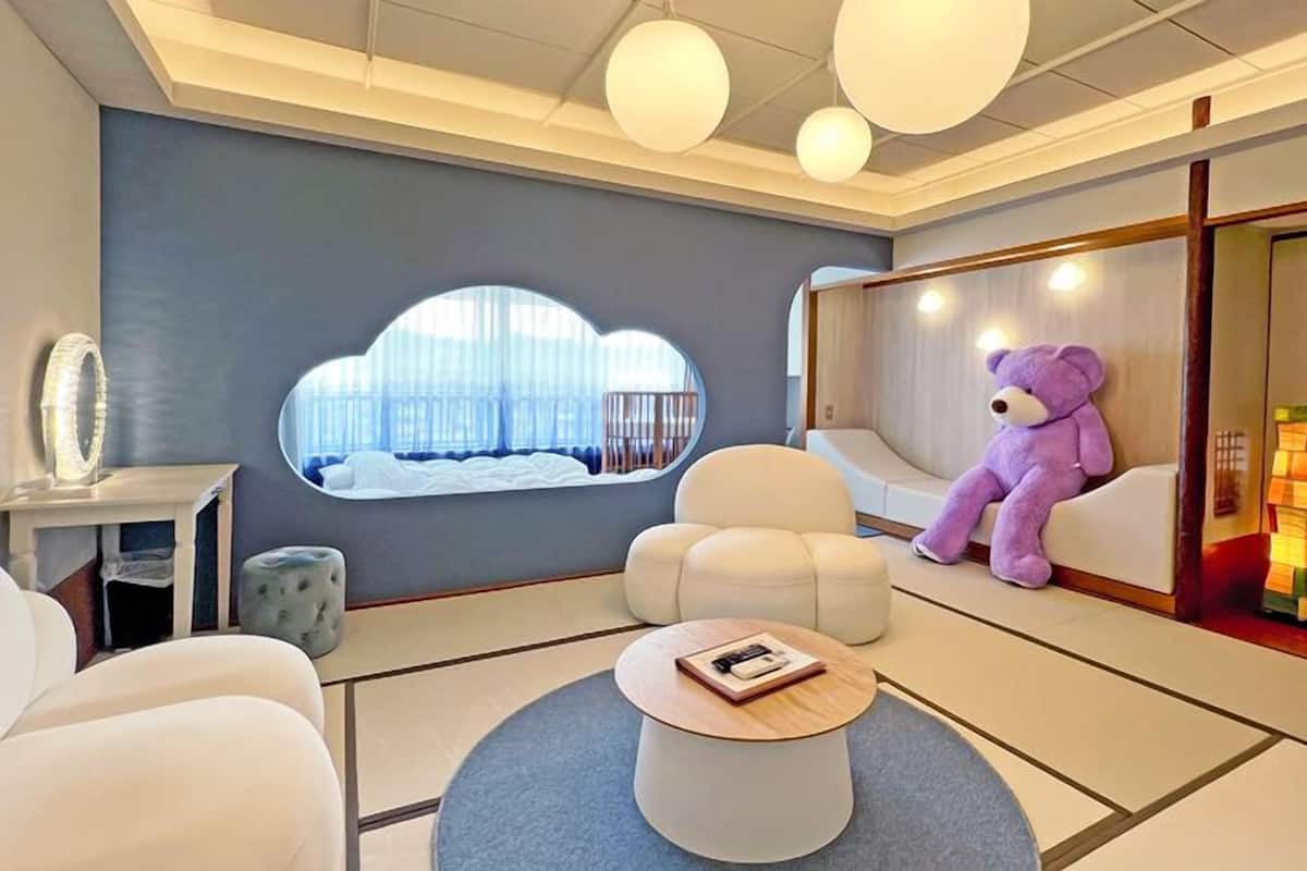 Une chambre d’hôtel moderne avec une grande fenêtre, un canapé moelleux, une table basse ronde et un ours en peluche.