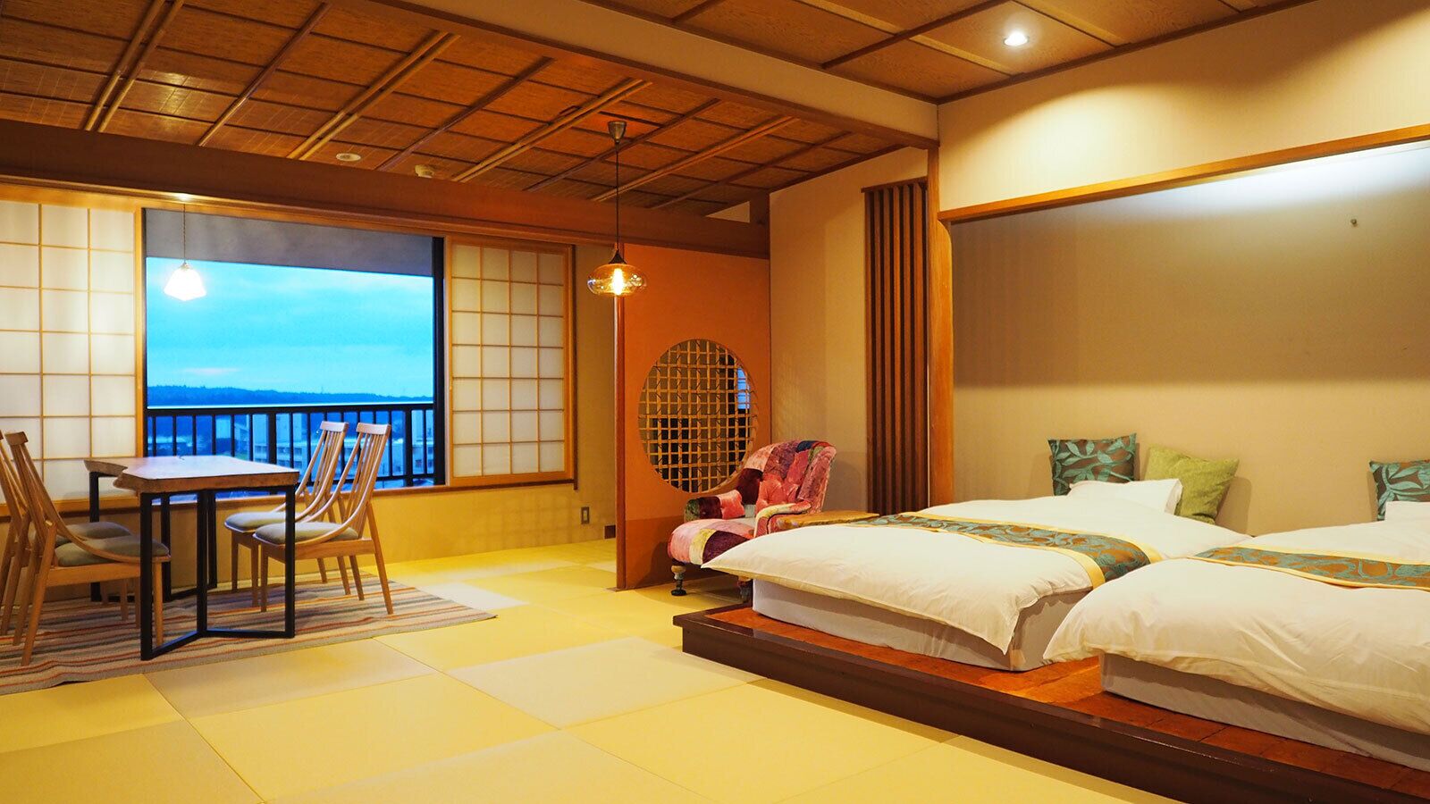 Una habitación con suelo de tatami, una cama de estilo japonés tradicional, una mesa con sillas y vistas al mar a través de un ventanal grande.