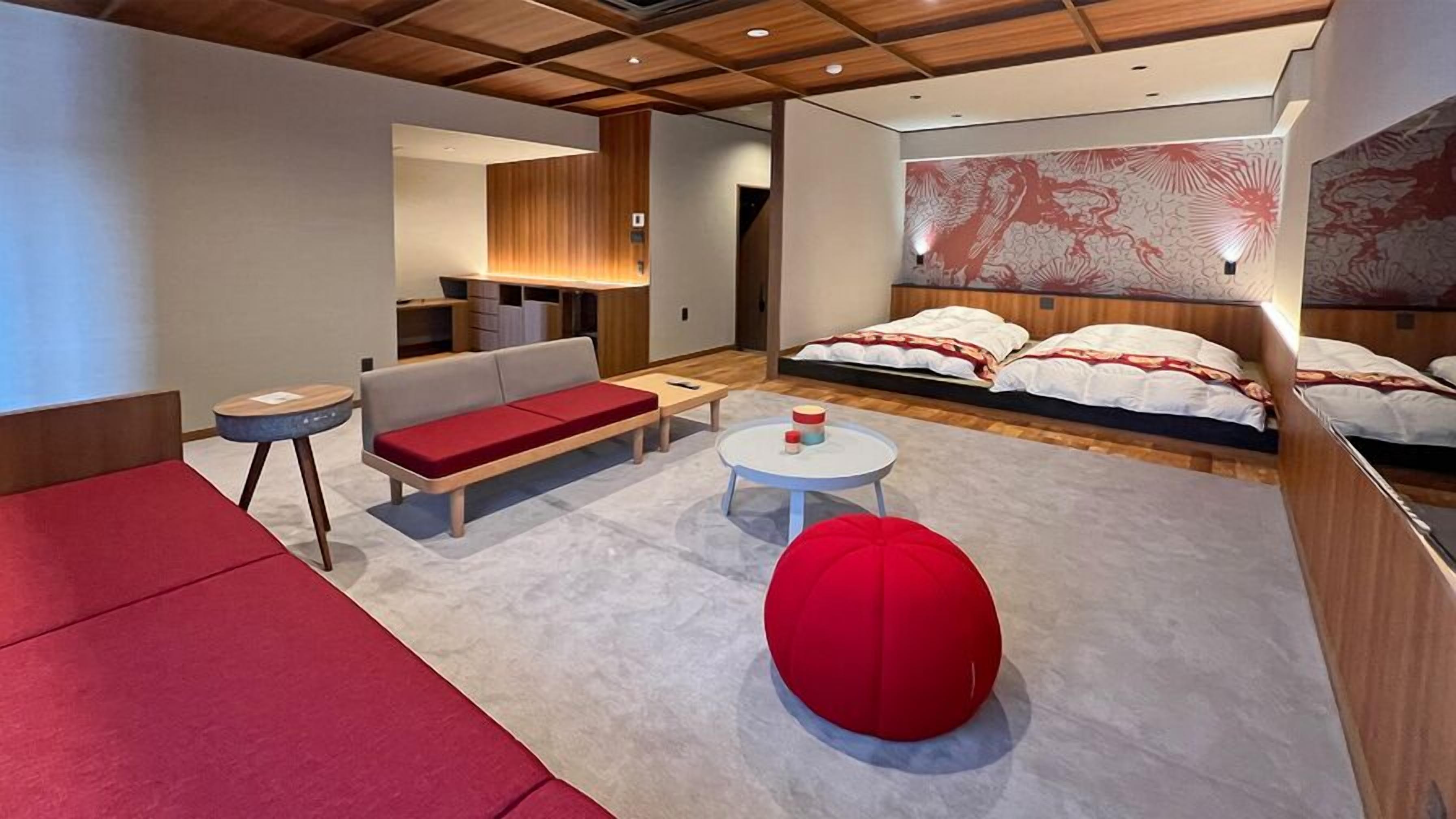 Una habitación de hotel moderna con sofá, una mesa redonda, un otoman rojo y una cama con un gran mural en la pared.