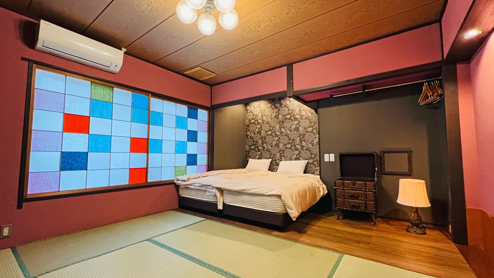 Una habitación con una cama, aire acondicionado de pared, una ventana con un motivo de cuadrícula de colores y una lámpara tradicional al estilo japonés.