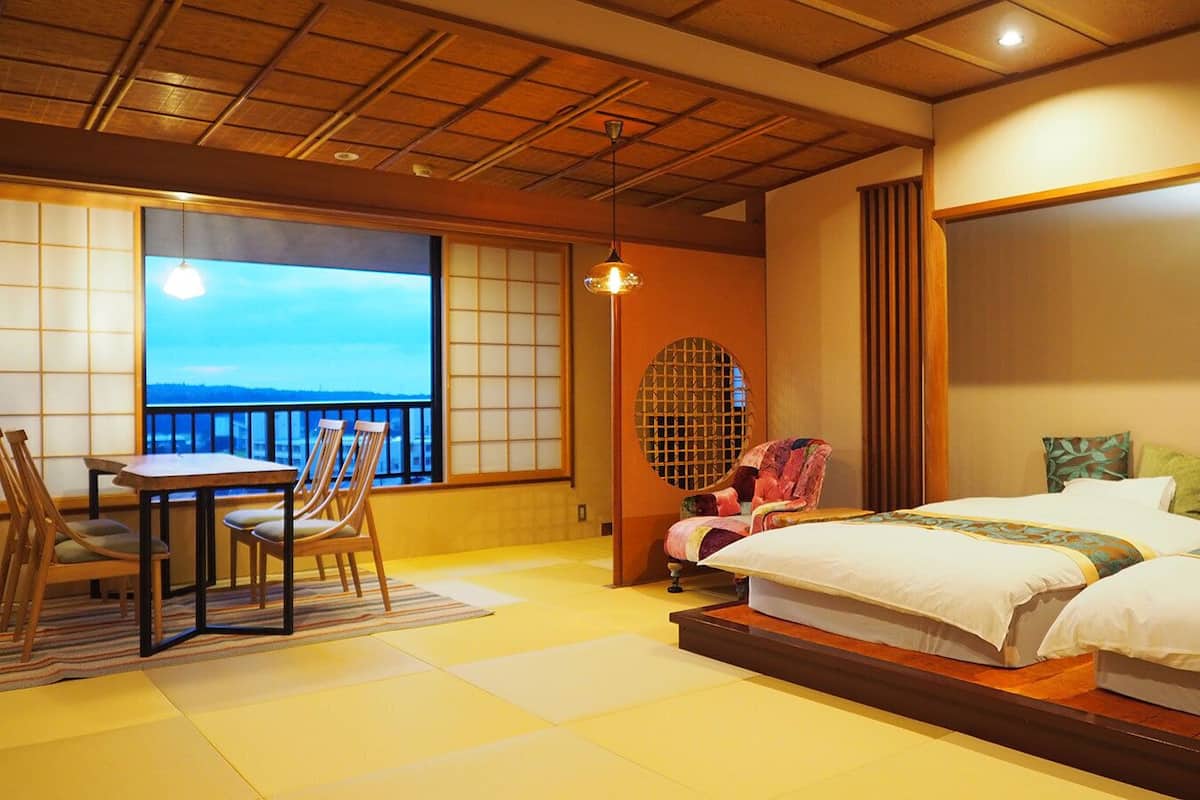 Une pièce de style japonais traditionnel, avec un sol recouvert de tatamis, une table basse entourée de chaises, un lit avec deux oreillers et une vue sur la mer par une grande fenêtre.