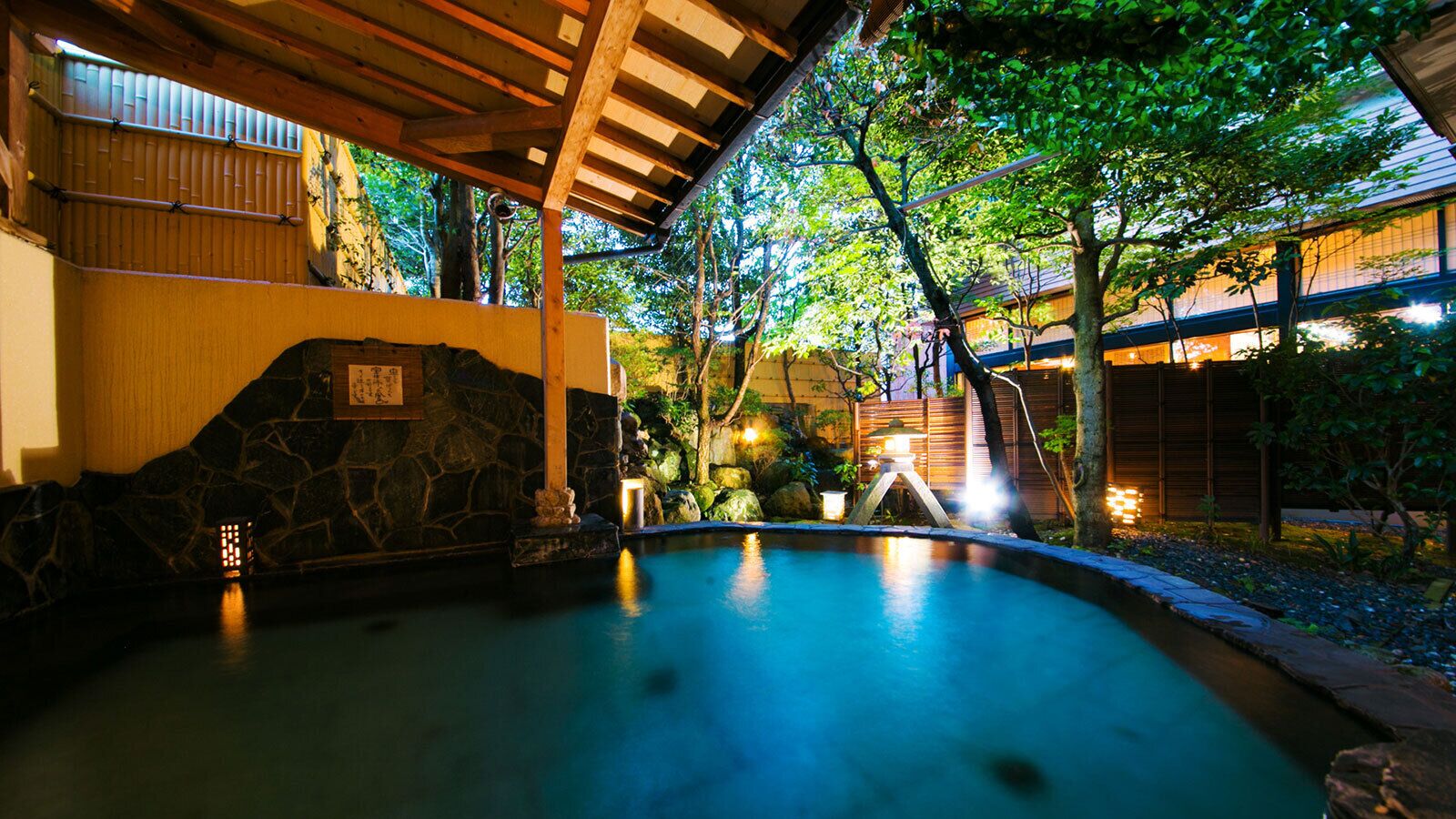 A tranquil indoor swimming pool with stone walls and a wooden ceiling, surrounded by lush greenery and traditional Japanese architecture.