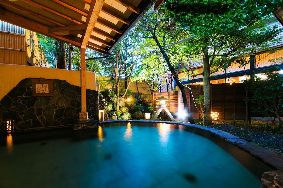 Une piscine intérieure paisible, avec des murs en pierre et un plafond en bois, entourée d’une végétation luxuriante et d’une architecture japonaise traditionnelle.