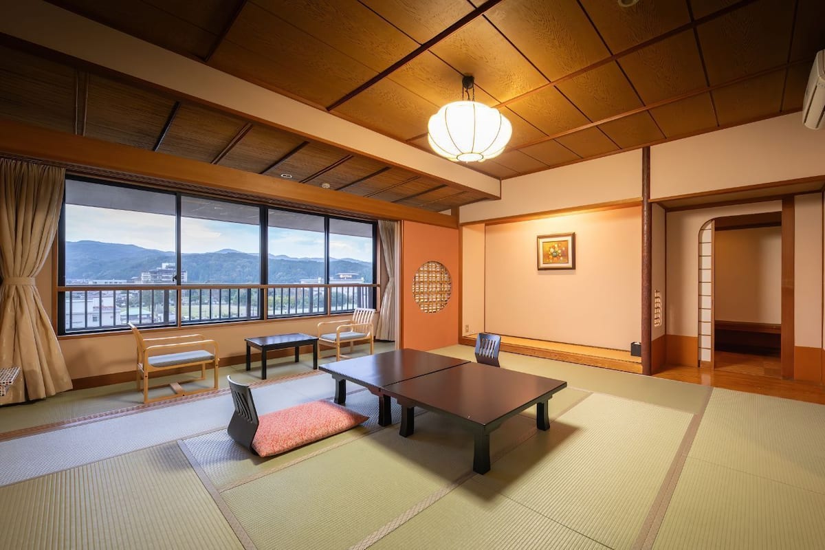 Une pièce de style japonais traditionnel, avec un sol recouvert de tatamis, une table basse, des chaises, une vue dégagée sur l’extérieur grâce à de grandes fenêtres et un ventilateur de plafond.