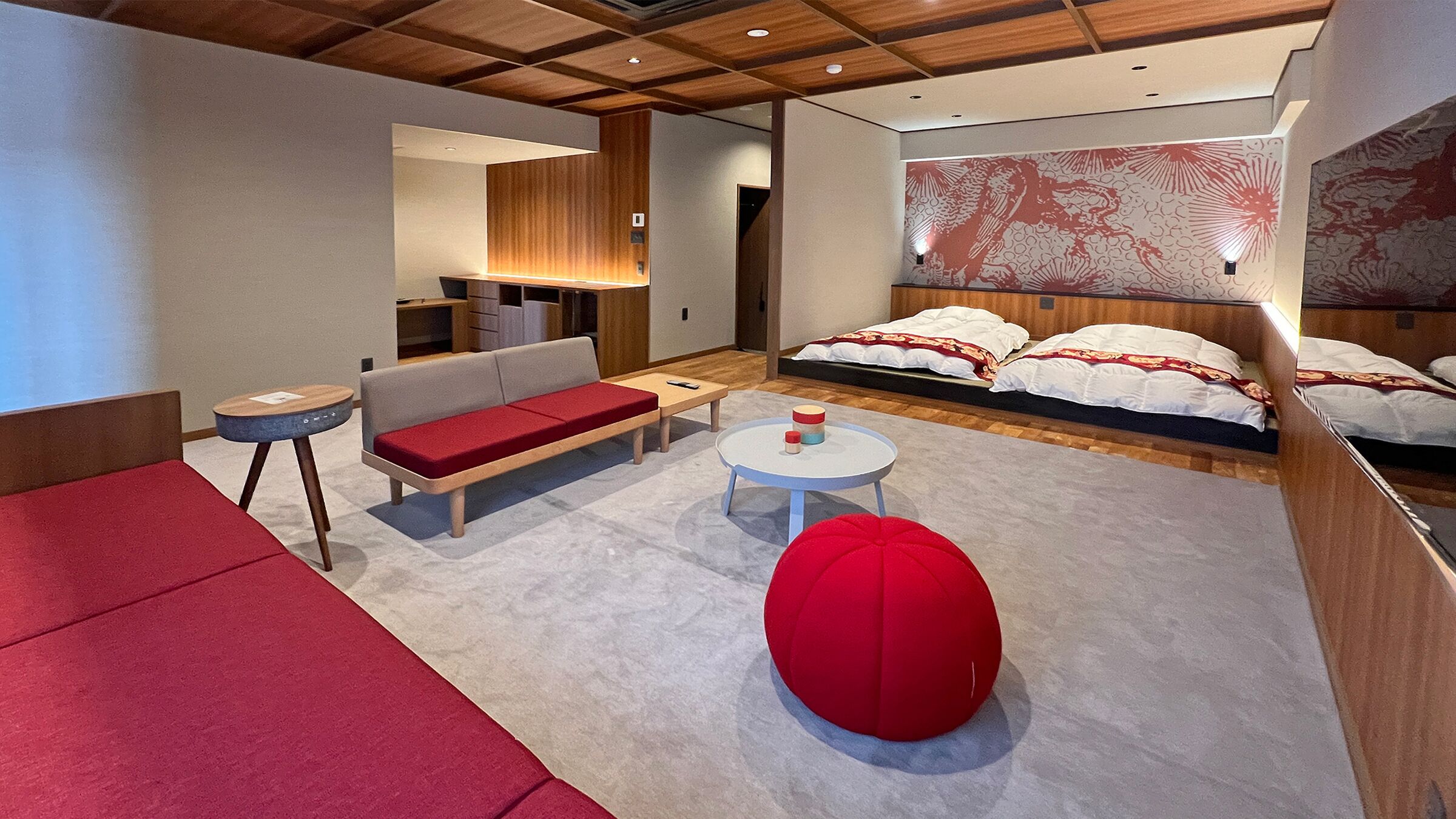 Una habitación de hotel moderna con sofá, una mesa redonda, un sillón de peluche rojo, una cama con ropa de cama estampada y un mural grande en la pared.