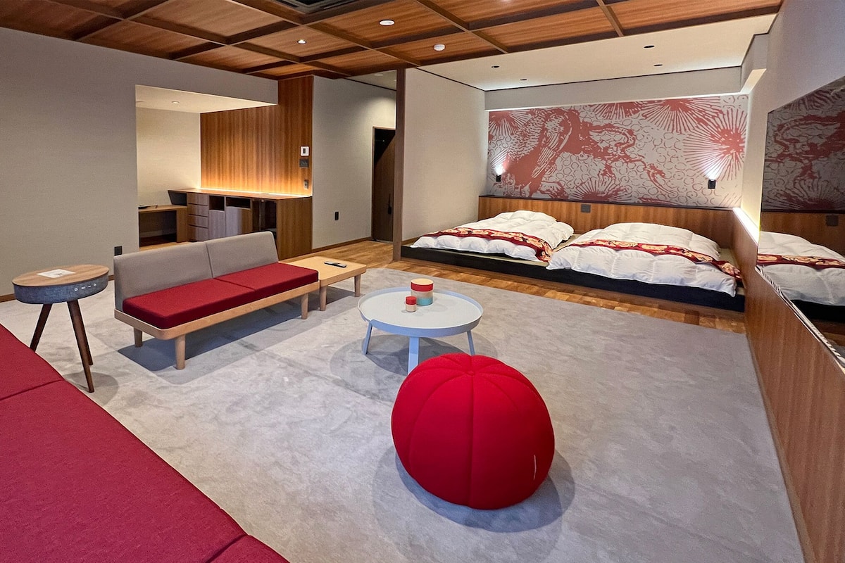 Une chambre d’hôtel moderne avec un canapé, une table ronde, un pouf rouge, un lit avec une literie à motifs et une grande fresque murale.